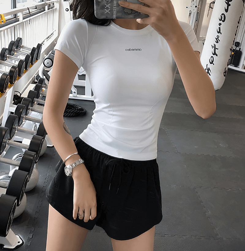 Woman wearing a white breathable fitness t-shirt and black shorts in a gym, showcasing comfortable and stylish workout apparel.